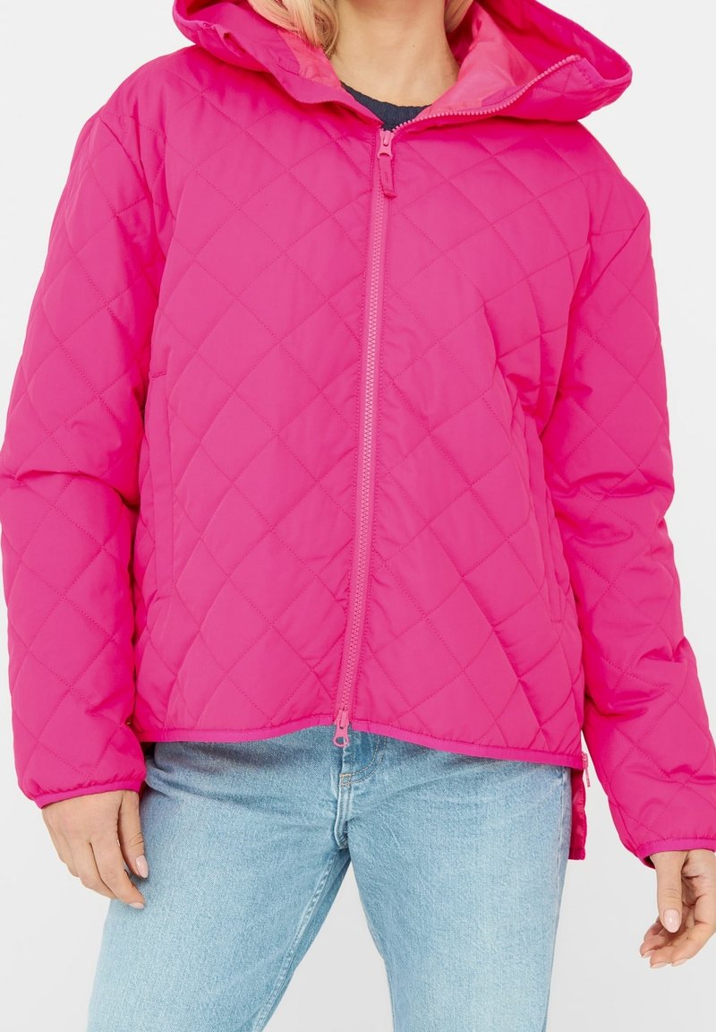 Femme portant une veste matelassée rose vif avec capuche, fermeture éclair à l'avant, associée à un jean bleu clair sur fond blanc.