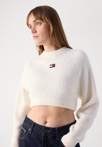 White cropped sweater with ribbed texture and long sleeves, featuring a small red, white, and blue logo on the front. Worn with dark jeans.