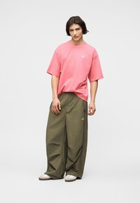 Νεαρός άνδρας με μακριά σκούρα μαλλιά ως τον ώμο, φορώντας μια ευρεία ροζ μπλούζα, υπερμεγέθη χακί παντελόνια και λευκά αθλητικά παπούτσια, στέκεται πάνω σε έναν απλό φόντο.