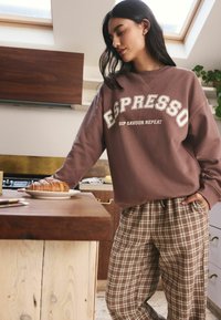 Brauner Sweatshirt mit dem Aufdruck "ESPRESSO" und cremefarbenen Akzenten, kombiniert mit karierten Hosen in Brauntönen. Holztresen mit einem Croissant auf einem Teller.
