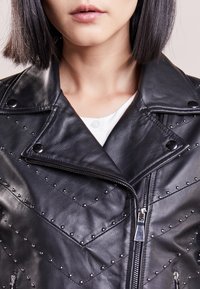 Zwarte leren jas met studs, diagonale stikpatronen en een asymmetrische ritssluiting. Gladde textuur en brede revers.