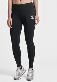 Osoba ubrana w czarne legginsy Hummel i szaro-białe buty sportowe stoi na tle gładkiej białej ściany.