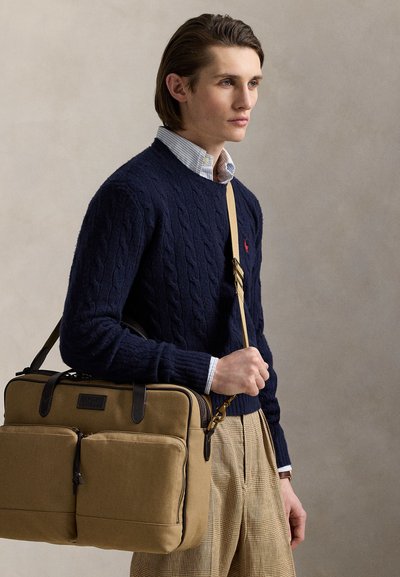 Young man wearing navy cable-knit sweater and plaid trousers holds tan messenger bag with two front pockets and shoulder strap.