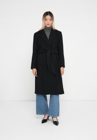 Lungo cappotto nero con design avvolgente, cintura a cravatta in vita, colletto a punte e grandi revers. Indossato sopra un dolcevita grigio e jeans svasati.