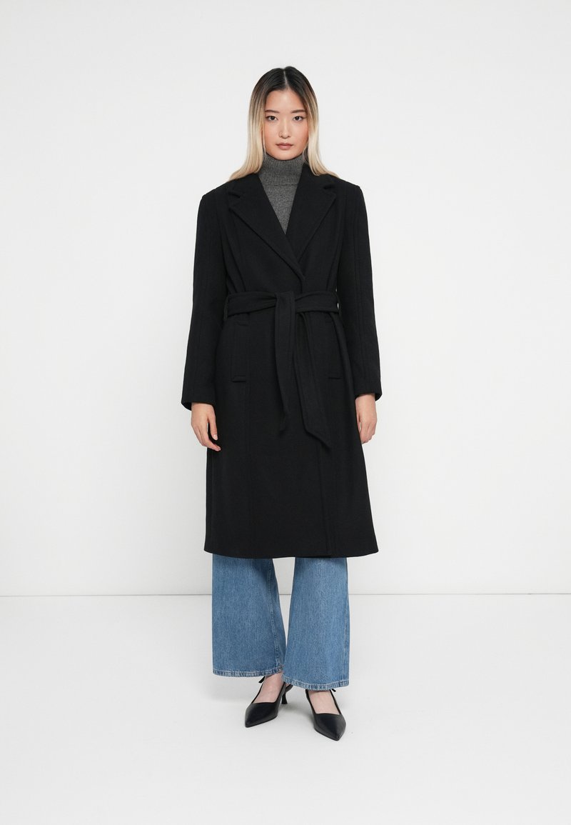 Lungo cappotto nero con design avvolgente, cintura a cravatta in vita, colletto a punte e grandi revers. Indossato sopra un dolcevita grigio e jeans svasati.
