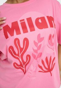 Camiseta de algodón rosa con el texto "Milan" en rojo intenso, y varios diseños de hojas en tonos más oscuros de rojo y rosa en la parte frontal.