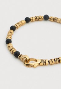 Βραχιόλι με χάντρες σε χρυσαφί απόχρωση που διαθέτει υφασμένες χρυσαφένιες και λείες μαύρες χάντρες, με κούμπωμα με άγκιστρο.