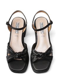 Sandalo in pelle nera con punta quadrata e dettaglio a fiocco annodato. Presenta una fascia alla caviglia e un tacco wedge testurizzato. Rivestimento interno liscio.