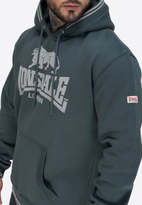 Sudadera verde con logo gris, detalle de cordón y bolsillo tipo canguro. Presenta acentos a rayas en la capucha y parche del logo de Lonsdale en la manga.