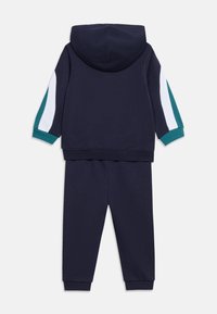 Σετ μπλε σκούρας φούτερ με κουκούλα και παντελόνι με ρίγες σε πετρόλ και λευκό στα μανίκια, σε οριζόντια προβολή από πίσω.