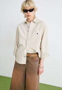 Beige button-up shirt with rolled sleeves, front pocket, and logo patch; paired with brown wide-leg jeans and a patterned brown belt. Black sunglasses.