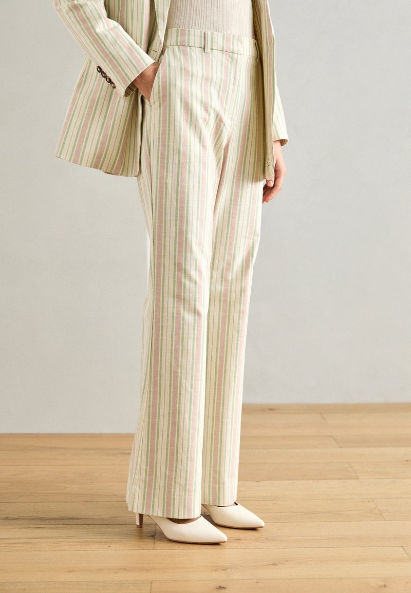 Personne portant un pantalon de costume rayé vertical beige et vert et un blazer assorti, associé à des talons blancs à bouts pointus, debout sur un sol en bois.