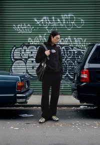 Sort hættetrøje med frontprint, sorte bukser med vidde, mønstret taske og unikke gule og sorte sneakers. Urbant baggrund med graffiti.