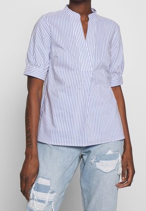 Osoba ubrana w bluzkę w pionowe niebiesko-białe paski z rękawami do łokcia oraz jasnoniebieskie podarte dżinsy.