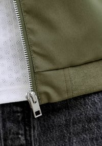 Õlgrohelise värvusega jakk, millel on tõmblukk, soonilised mansetid ja tekstuurse sisemise voodriga, esitleb tumedal taustal.