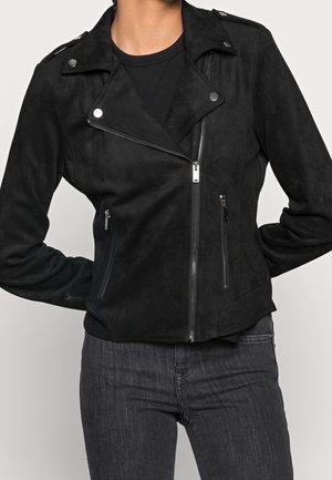 Woman wearing a black suede moto jacket with silver zipper details and buttons, paired with dark gray jeans and a black shirt.