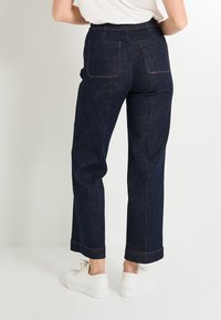 Pantalones de mezclilla oscuro, de tiro alto y pernera recta, con dos bolsillos traseros y costuras visibles; combinados con zapatillas blancas.