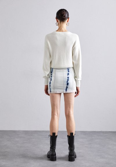 Pull en maille côtelée blanc, jupe en denim avec des accents en tie-dye bleu, et bottes en cuir noires à hauteur de genoux avec des talons épais. Modèle vu de dos.