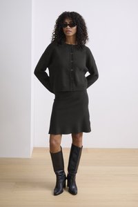 Tmavě pletený cardigan s knoflíky, spárovaný s úzkou tmavou sukní. Model má na sobě vysoké černé kožené boty. Hladká textury a minimalistický design.