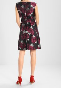 Černé šaty s květinovým vzorem v burgundské a stříbrné barvě, přiléhavý střih, po kolena, bez rukávů, v kombinaci s červenými podpatky.