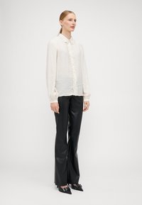 Blouse claire à manches longues avec une finition texturée et un plastron décoratif à l'avant, assortie de pantalons noirs brillants et de ballerines pointues.