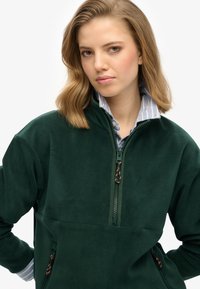 Donkergroene fleece trui met een hoge kraag, ritssluiting aan de voorkant en twee zijzakken. Binnenkant heeft een lichtblauwe gestreepte overhemdkraag.