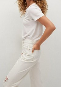 Bílý tričko s krátkým rukávem v kombinaci s džínami s vysokým pasem ve světlé barvě, které mají oprané akcenty na levém stehně.