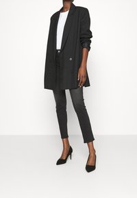 Musta kaksirivinen bleiseri, jossa suuret napit, valkoisen paidan päällä, yhdistetty kapeisiin mustiin farkkuihin ja teräviin mustiin korkokenkiin, neutraalilla taustalla.