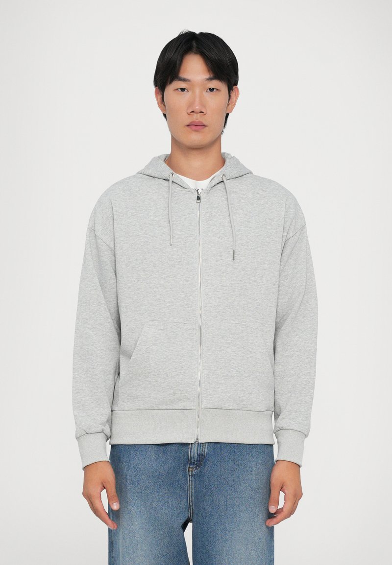 Sudadera gris con cremallera y capucha con cordón, con bolsillos frontales, que presenta una textura suave y puños y dobladillo acanalados. Usada con jeans azules.