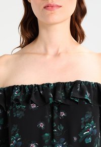 Black off-the-shoulder top with ruffled neckline, featuring green and white floral patterns against a dark background. Smooth fabric texture.