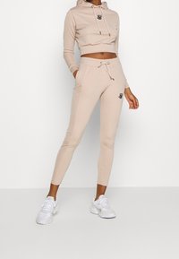 Beige verkürzter Hoodie mit Kordeln und Logo, kombiniert mit beigen schmal zulaufenden Jogginghosen mit elastischem Bund und seitlichen Taschen.