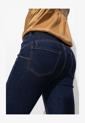 Jeans en denim bleu foncé au design ajusté, cinq poches et des coutures orange contrastantes sur les coutures. Ceinture visible.
