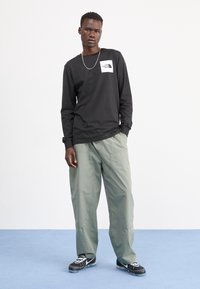 Schwarzes langärmliges Shirt mit weißem Logo, leichte grüne Cargo-Hosen und schwarze Sportschuhe mit weißen Akzenten, stehend auf einem blauen Teppich.