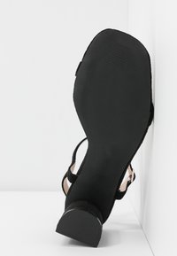 Sandale en daim noir avec une semelle en caoutchouc noir texturé. Dotée d'un talon ouvert et d'une bride à la cheville, avec un léger talon bloc et des accents lisses.