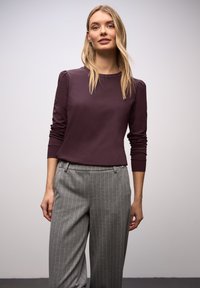 Dunkelrotes Langarmshirt mit rundem Ausschnitt und gerafftem Schulterdetail, kombiniert mit grauen, feingestreiften Hosen mit seitlichen Taschen.