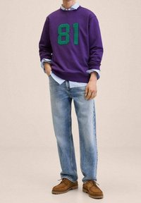 Sudadera morada con el estampado verde "81", superpuesta a una camisa de color azul claro con cuello. Combinada con vaqueros azules desgastados y zapatos marrones.