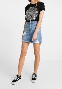 Svart t-shirt med silver mandala grafik, kombinerad med en ljusblå denim-minikjol med fransad nederkant och slitet utseende, bärs med svarta sneakers.