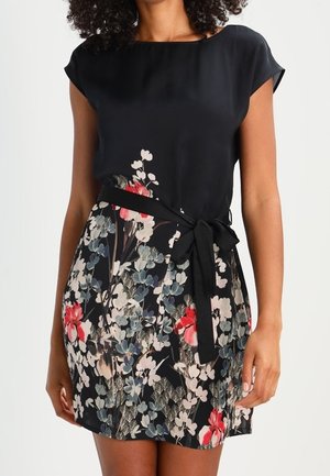 Kobieta w czarnej sukience z kwiecistym wzorzystym dołem i czarną kokardą zawiązaną w talii, stojąca na jednolitym tle.