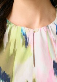 Blouse multicolore avec un col rond, une petite fermeture à bouton et une fente verticale. Le tissu a une texture douce et lisse avec des motifs abstraits.