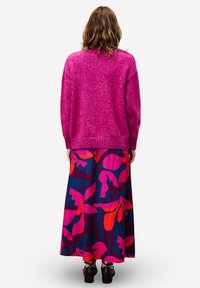 Vrouw met golvend bruin haar, gekleed in een felroze sweater en een lange bloemenrok met roze, rode en marineblauwe patronen, staande met haar rug naar de kijker gekeerd.