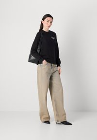 Kvinne med langt mørkt hår står selvsikkert, iført en svart hettegenser og lyse brede jeans, mens hun holder en svart håndveske.