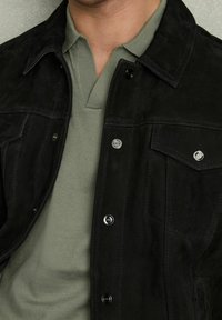 Mężczyzna w czarnej zamszowej kurtce na jasnzie zielonej koszuli z kołnierzykiem, ukazujący górną część klatki piersiowej i szyję.