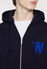 Navy zip-up hoodie with a drawstring hood, featuring a blue embroidered letter on the chest and a silver zipper. Smooth fabric texture.