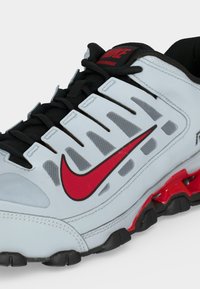 Λευκό και μαύρο αθλητικό παπούτσι Nike με κόκκινο λογότυπο και λεπτομέρειες, με διχτυωτό αερισμό και υφασμένη σόλα για αθλητική χρήση.