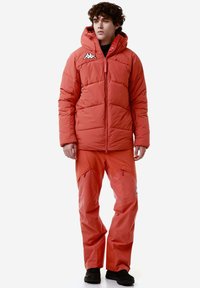 Giacca arancione puffa con cappuccio e motivo cucito, abbinata a pantaloni sportivi arancioni coordinati. Presenta il logo Kappa su entrambi i capi.