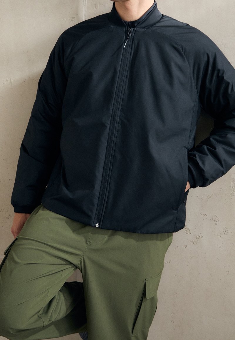 Giacca nera con zip e colletto alto, dal design imbottito. Indossata sopra pantaloni cargo verde oliva con tasche laterali e una vestibilità comoda.