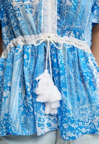 Blusa blu a fantasia con disegni intricati, bordo in pizzo bianco e vita con lacci e pon pon bianchi. Tessuto morbido e leggero.