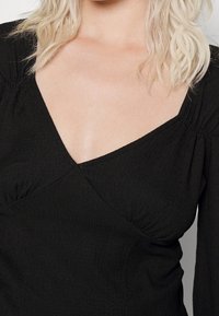 Svart texturerad topp med djup V-ringning och draperad detaljering på ärmarna, som har en figurnära silhuett och mjuk kvalitet.