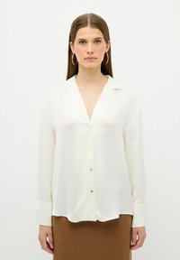 Blusa bianca in satin con scollatura a V, maniche lunghe, chiusura con bottoni e polsi con polsini, abbinata a una gonna marrone. Design minimalista e texture liscia.