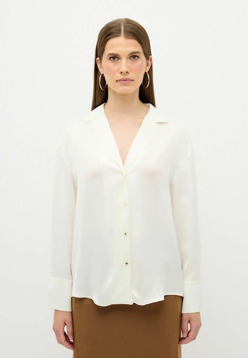 Blusa bianca in satin con scollatura a V, maniche lunghe, chiusura con bottoni e polsi con polsini, abbinata a una gonna marrone. Design minimalista e texture liscia.